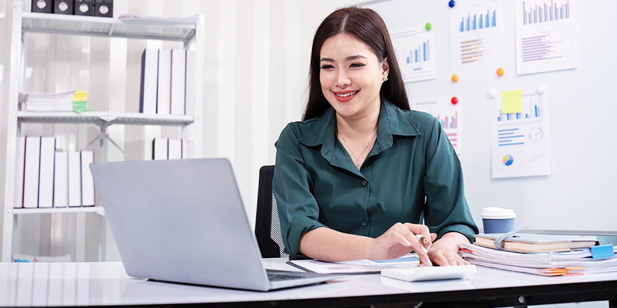 photo of a woman wearing a green blouse, sitting at her desk, smiling, looking at her laptop and punching numbers into a calculator