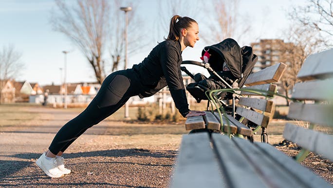 A woman does some postpartum pelvic floor exercises in a park. She is leaning on a bench and her baby is in a pram behind her. 