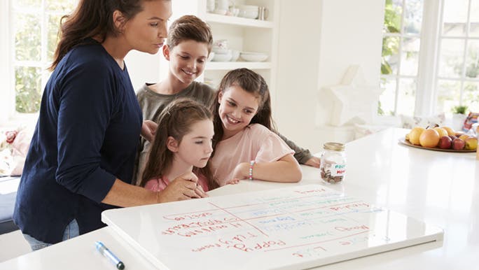 A mum shows her three primary aged kids a routine chart she has made on a whiteboard.