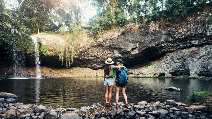Two people are standing at a waterhole in the Australian bush, they have been hiking and have their arms around each other. 