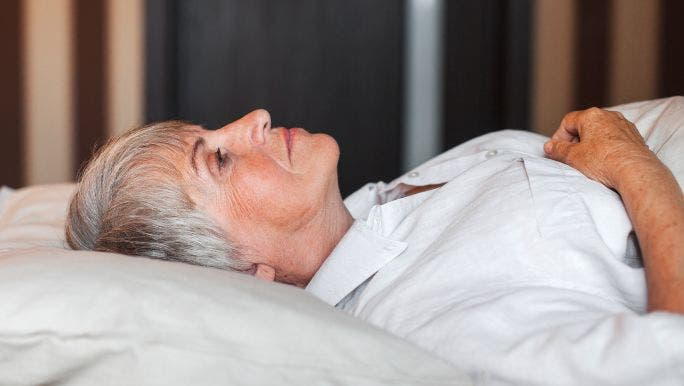 A senior woman wearing white pyjamas is lying in bed looking up with a neutral expression