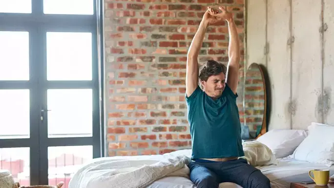 Man in blue pyjamas sitting up and stretching in bed in the morning
