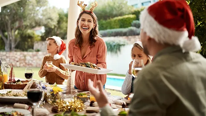 Woman is smiling carrying a tray she is about to set down on the outdoor dining table. She is at a Christmas family lunch and there is a festive spread of food and drink on the table.