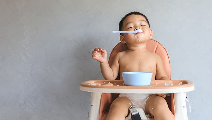 A very satisfied looking baby is holding a spoon in their mouth and leaning back with their eyes closed. They are sitting in a high chair in front of a grey wall. 