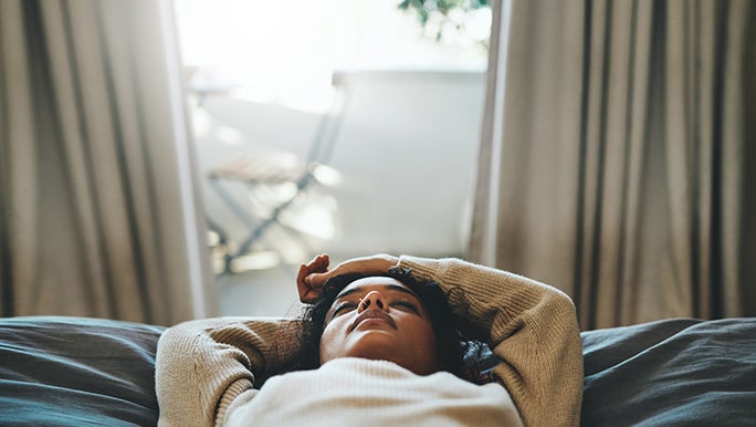 A lady is laying on a bed with her eyes closed and her arms above her head. There is a window behind her with open curtains. She doesn’t seem to be asleep though.
