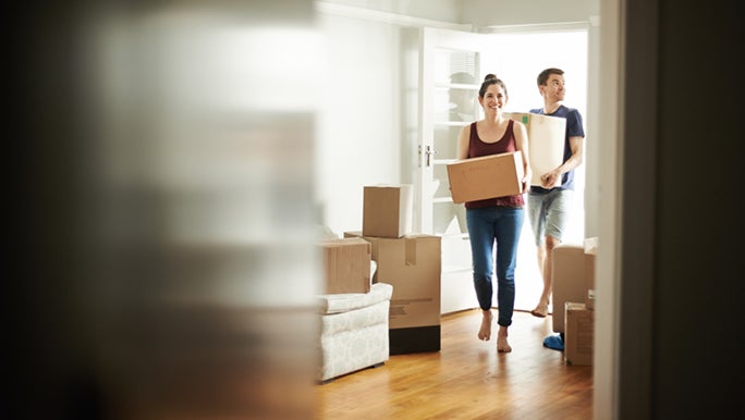 A couple carrying boxes are walking through a doorway into their new home. They followed a bunch of moving house tips and have had a stress-free move.