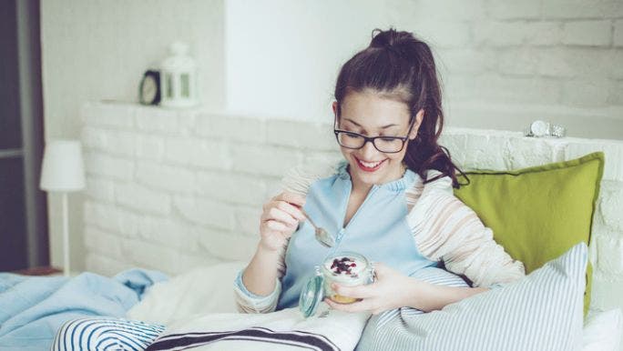 A young woman wearing glasses and smiling is sitting in bed surrounded by cushions eating yoghurt