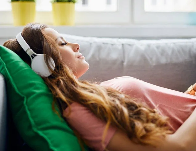 Young woman lying on the couch with a green cushion under her head. She is listening to headphones, relaxing with her eyes closed