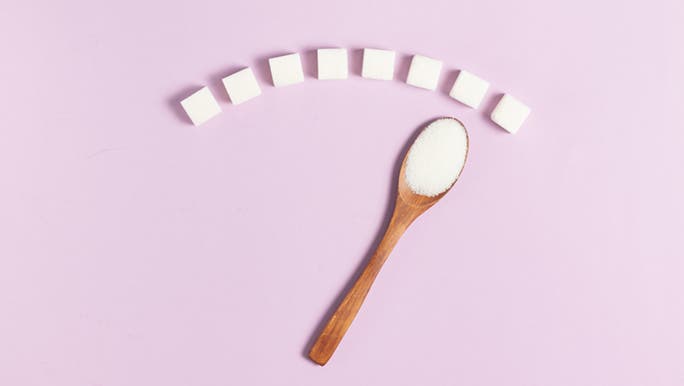 In front of a pink background, a wooden spoon full of sugar points at some sugar cubes to represent what sugar is.