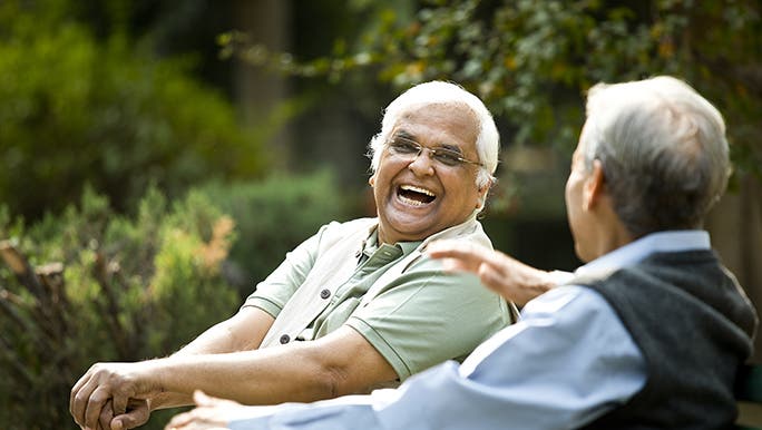 wo men sit on a park bench in the sun, chatting and laughing together. 