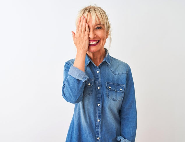 Middle age woman wearing casual denim shirt standing covering one eye with hand, confident smile on face and surprise emotion