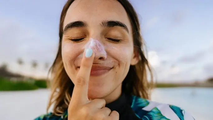 A woman in a sunshirt smiles as she puts sunscreen on her nose because she knows how sunscreen works. 