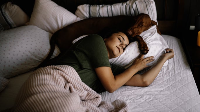 A lady sleeps soundly with her dog above her on the pillow, the is aware of how important sleep is to woman’s health.