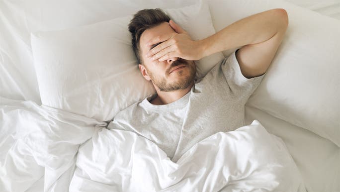 A dark-haired man wearing a T-shirt is lying in bed covering his eyes with his hand