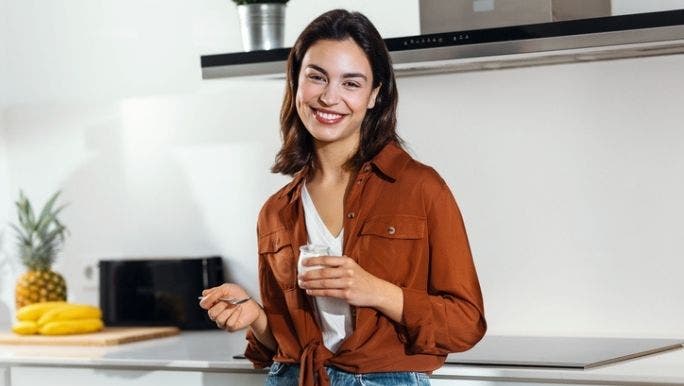 A young woman is smiling and leaning on a kitchen bench as she eats a tub of yoghurt