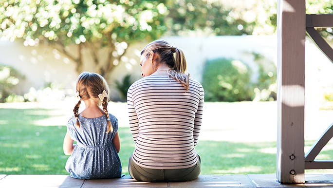 From behind, a mother and daughter are sitting on the step of a front porch. It’s a sunny day and they look to be having a chat. 