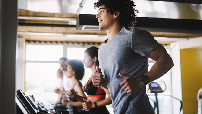 People in the gym running on treadmills with a younger, smiling man in the foreground who is working on ways to improve his cardio fitness.
