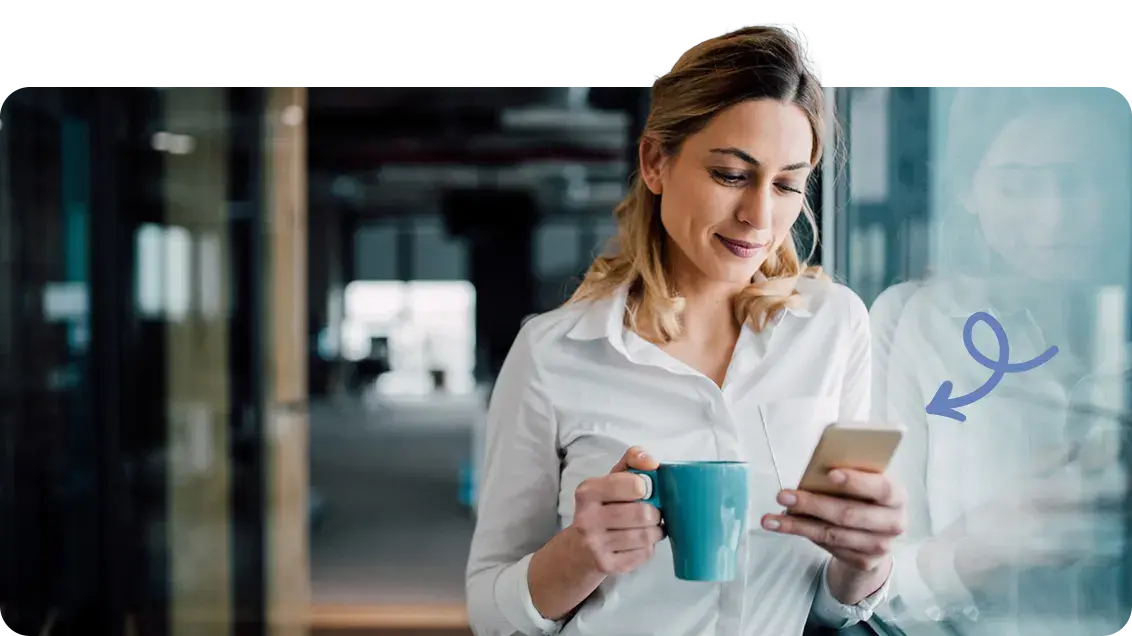 A woman stands next to a window. She is holding a blue coffee mug and a smart phone. She is looking down at her phone and smiling slightly. A blue graphic arrow points towards her phone.