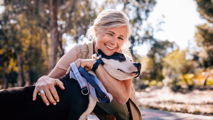 A lady is cuddling a dog in a park, they are meeting their minimum exercise requirements by going for a walk outdoors. 