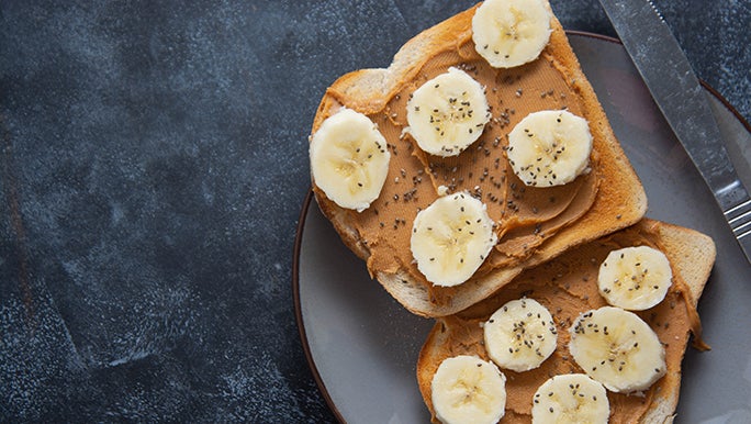 Two slices of peanut butter toast are on a plate, they have sliced banana and chia seeds on them.