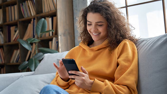 Smiling woman in orange hoodie using a habit tracker app on an iPhone. She is using a scheduling app to help her remember to take medication.