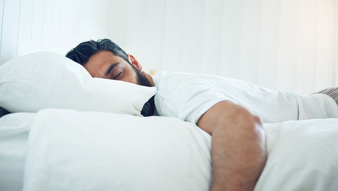 A dark-haired man is sleeping in a bed with white linen