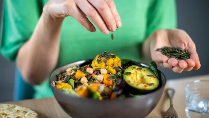 Woman in a green top adding pumpkin seeds to a healthy vegan meal in a bowl