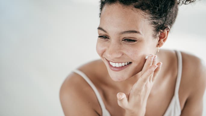 Smiling while applying moisturiser in the mirror, a woman is thinking about whether she has an oily or dry skin type. 