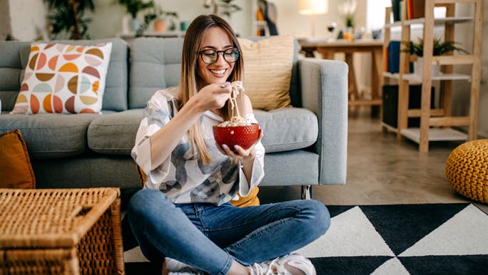 A young woman is sitting cross-legged on the floor eating a bowl of pasta