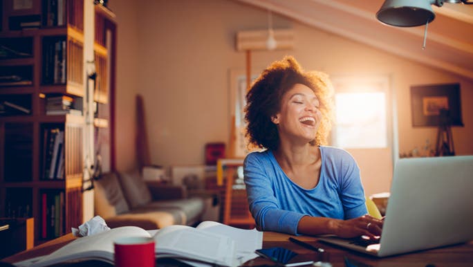 A woman in a home office throws back her head and laughs, it looks as though she is taking a study break to chat to a friend on her laptop.