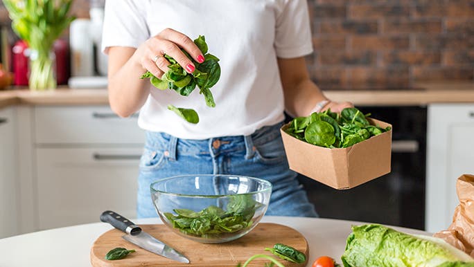 The torso of a lady is in the frame. She is standing in a kitchen and transferring raw vegetables from a cardboard box into a glass bowl. 