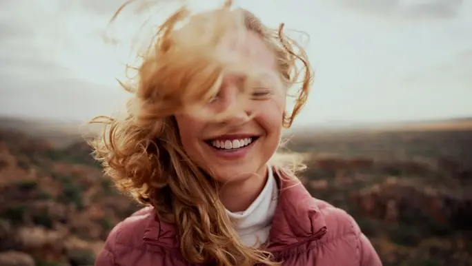 Smiling woman face partially covered with flying hair in the outdoors in a pink puffer jacket