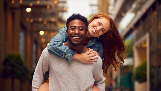 A dark-skinned young man is piggybacking his female partner who has long red hair as they walk down an alleyway smiling