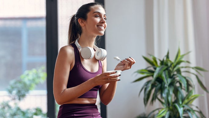 A young woman is wearing purple activewear and is smiling while she eats yoghurt