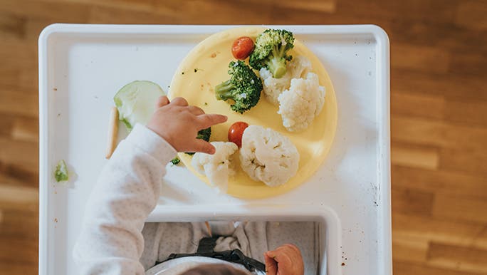 From above, a baby is eating in a white highchair. You can see the top of their head and little hands reaching for cauliflower and broccoli on a yellow plate. 