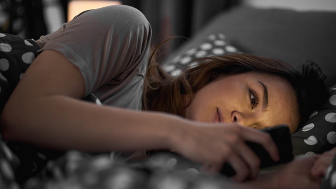 Woman is laying in bed looking at her phone, her sleep may be affected by blue light.
