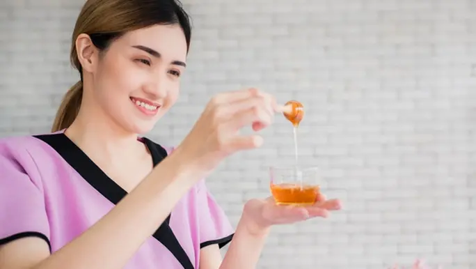Woman in a pink top drizzling honey into a glass bowl for her skincare routine