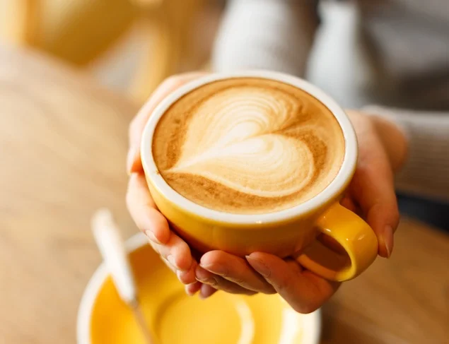 Freshly brewed flat white in a yellow coffee mug being cupped in a woman's hands