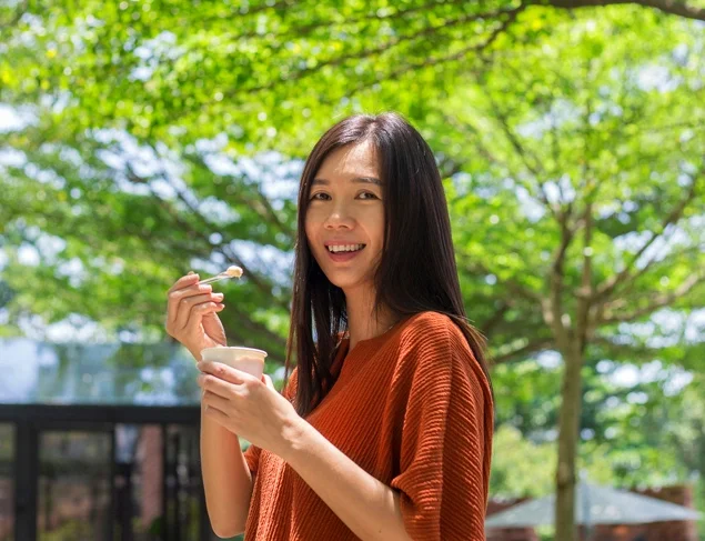 Smiling woman with long dark hair eating probiotic yoghurt in the park