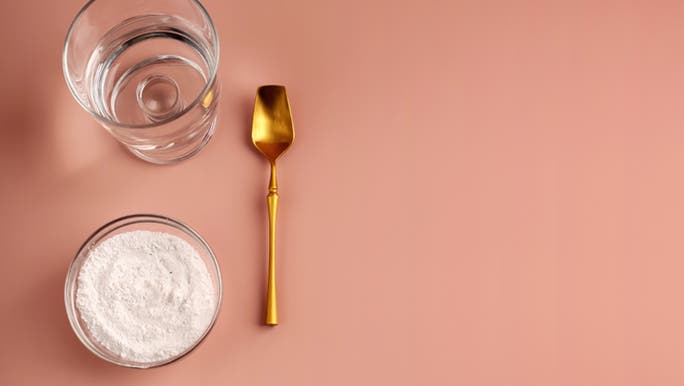 On a pink table, a glass of water sits with a gold spoon and a bowl of the best collagen powder in Australia.