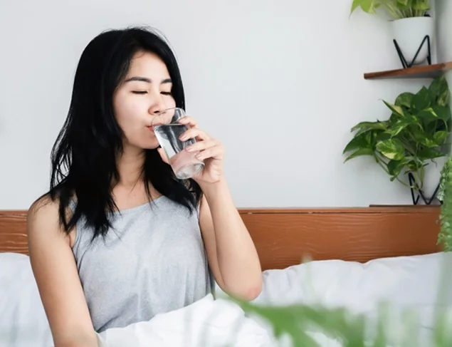 Woman with long black hair drinking a glass of water in bed first thing in the morning