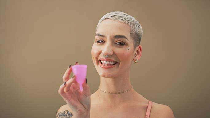 A smiling woman holds a pink menstrual cup which is an eco-period product that she uses.