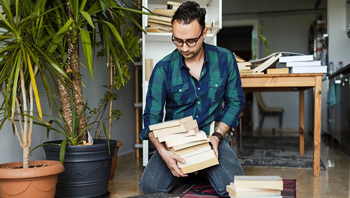 A man is sitting on the ground organising his book collection while stuck at home bored.