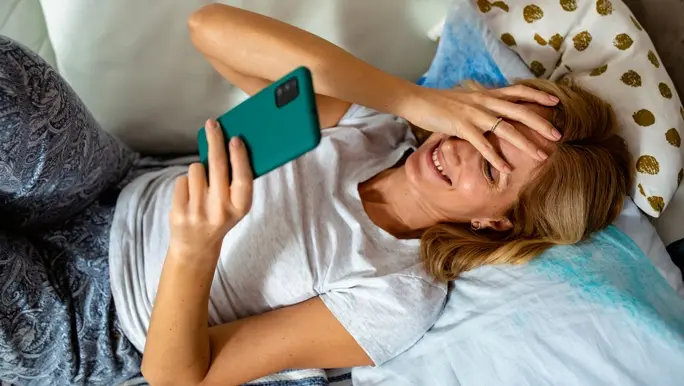 Woman using her smartphone as she lies down the length of the bed and smiling