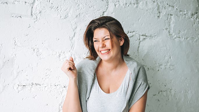 A lady is standing in front of a rendered grey wall, she is smiling ear to ear. 