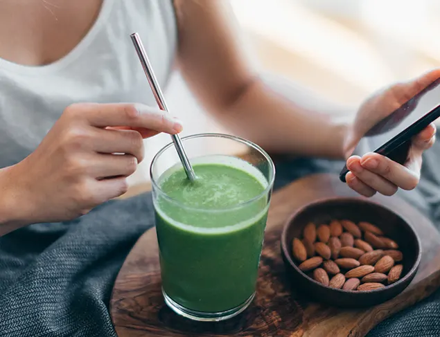 Woman (head cropped out) sits down at table while holding her phone in one hand ad holding her metal straw in the other. The straw is in a cup holding green powder mixed with water. Next to it is a dish filled with almonds.