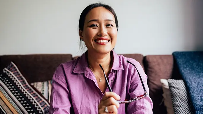 Woman wears a purple shirt and smiles towards the camera while holding her glasses in her hand.