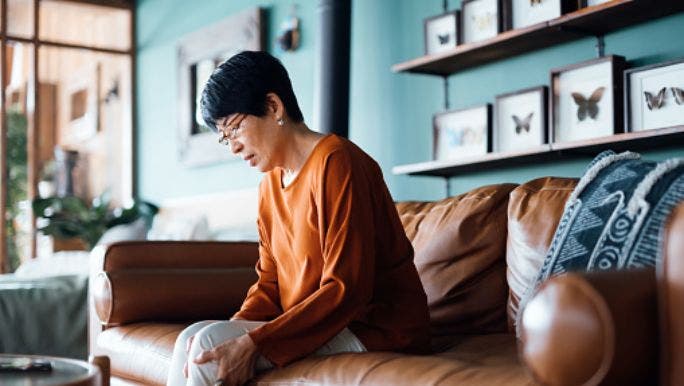 A senior Asian woman is sitting on a sofa and holding her knee