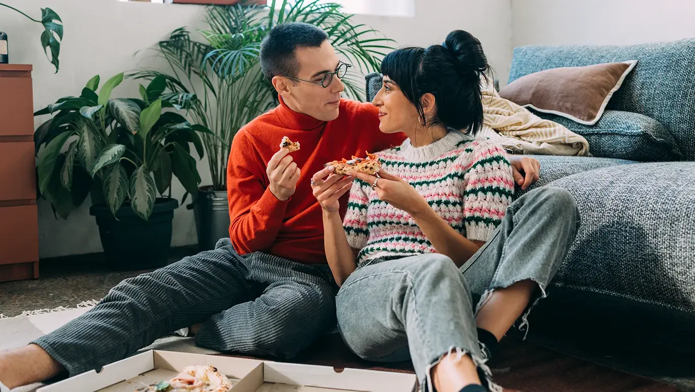 Couple sit on the floor eating pizza together in their living room.