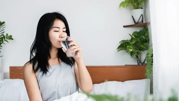 Woman with long black hair drinking a glass of water in bed first thing in the morning